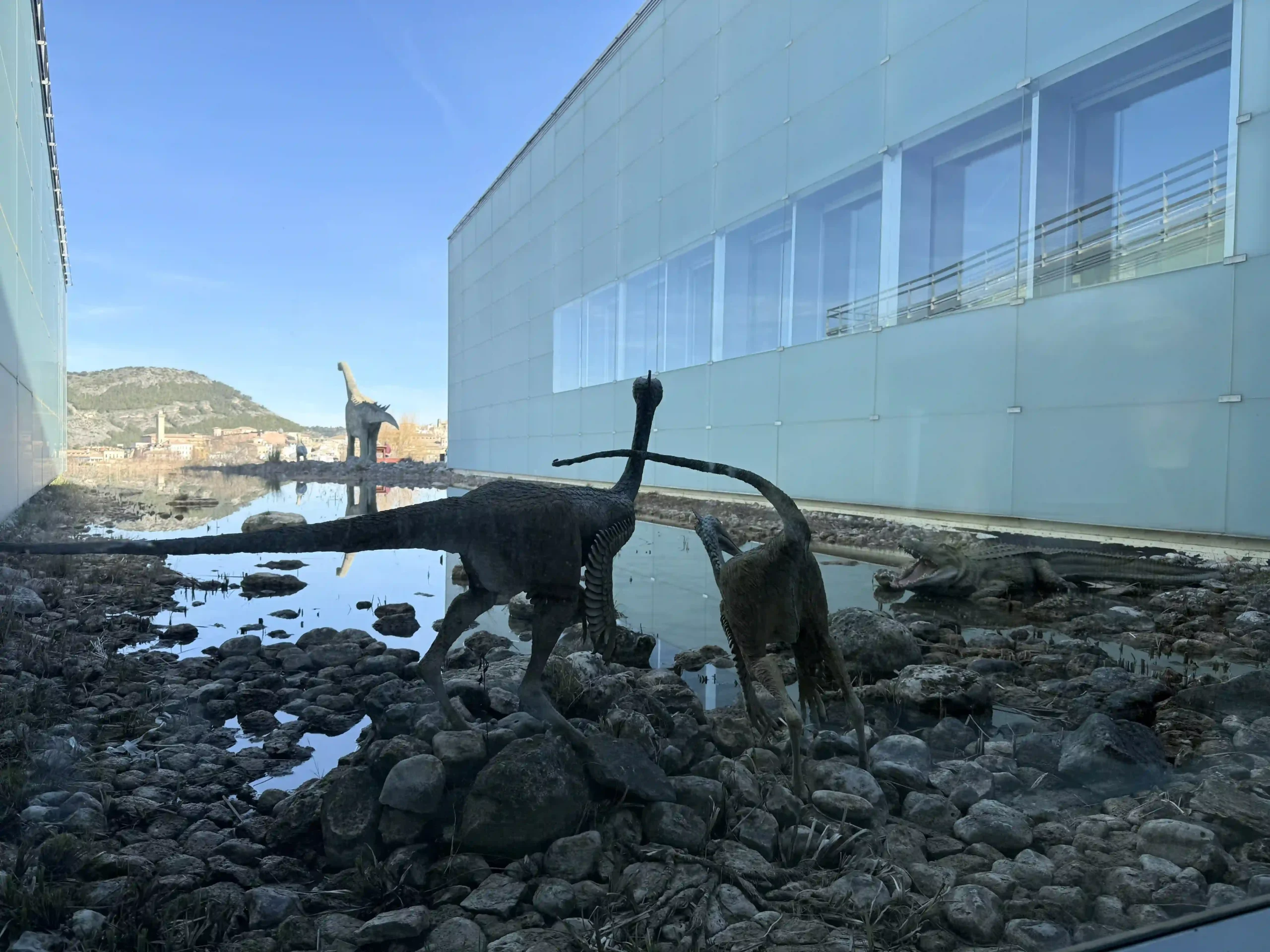 Reproducciones de dinosaurios en el estanque junto a la fachada acristalada del MUPA, con el casco de Cuenca al fondo. Foto propia La Maleta Inquieta