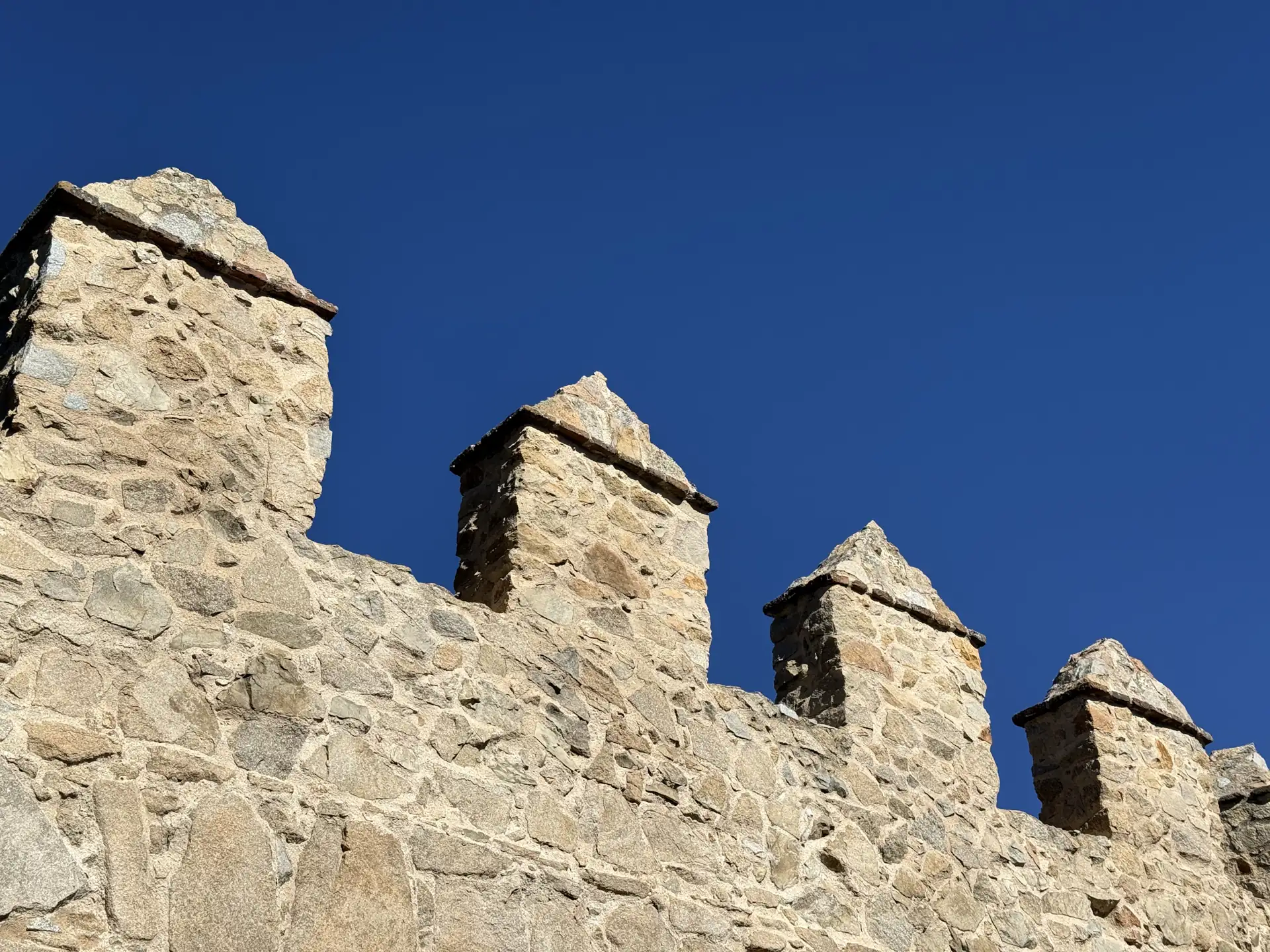 Almenas de granito rematadas en pirámide de la muralla de Ávila, vistas desde abajo contra un cielo azul intenso. Foto propia La Maleta Inquieta, septiembre 2026.