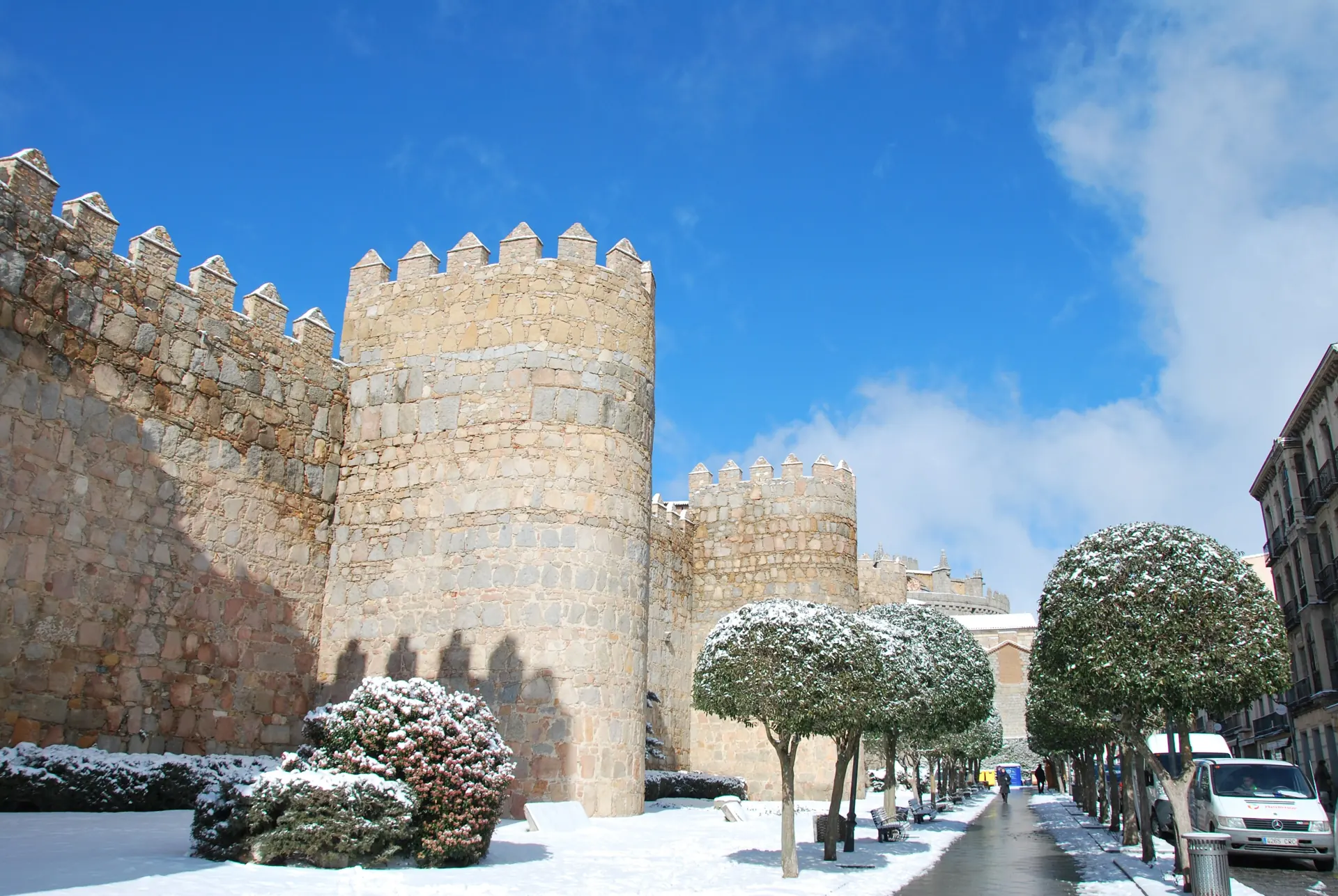 Cubos de la muralla de Ávila con nieve en el suelo y en los árboles del paseo, bajo un cielo azul de invierno