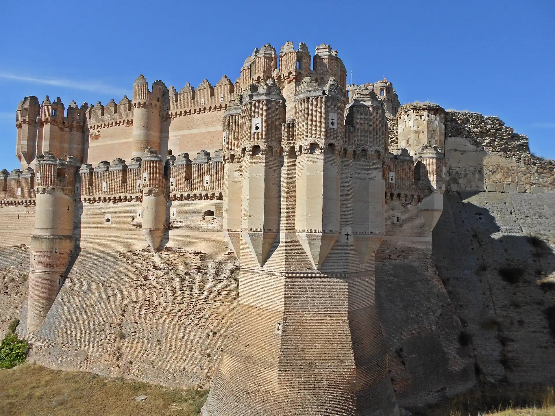 El castillo de Coca visto desde el borde del foso, con el muro de ladrillo y las torres poligonales.