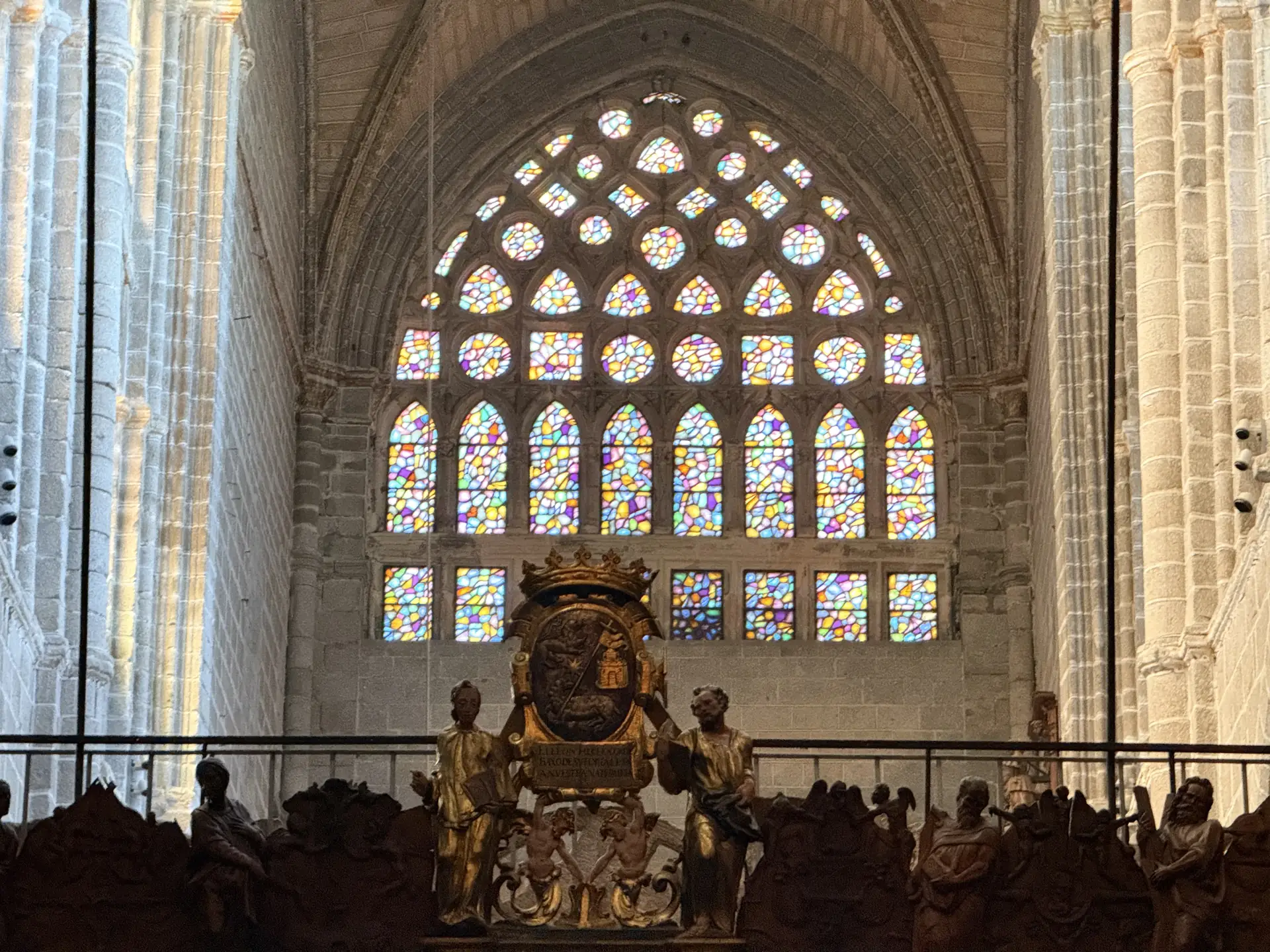 Remate dorado con escudo coronado de la sillería del coro de la catedral de Ávila ante un ventanal multicolor. Foto propia La Maleta Inquieta, septiembre 2026.