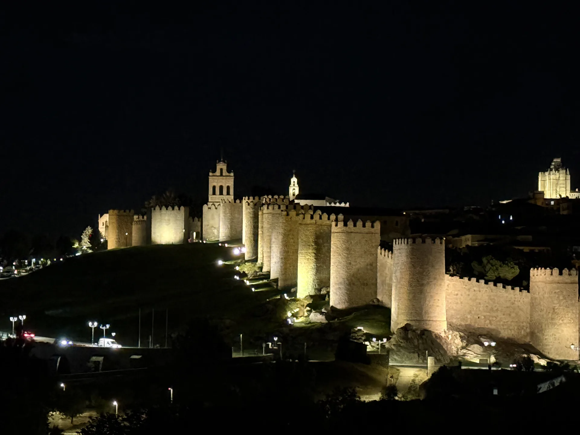 Cubos del lienzo norte de la muralla de Ávila iluminados de noche sobre la roca, con la espadaña del Carmen. Foto propia La Maleta Inquieta, septiembre 2026.