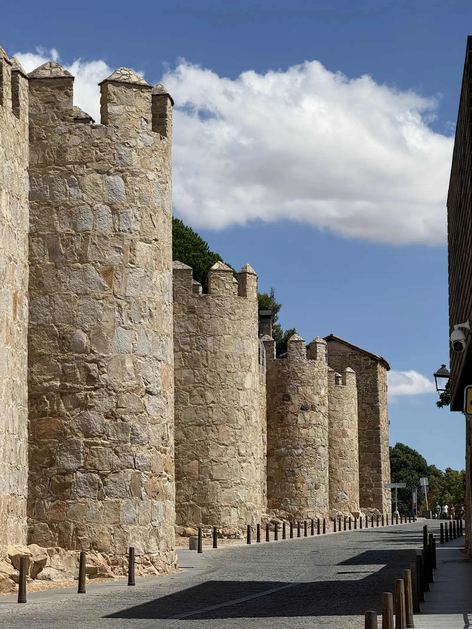 Hilera de cubos almenados de la muralla sur de Ávila junto a una calle empedrada con bolardos, en perspectiva. Foto propia La Maleta Inquieta, septiembre 2026.
