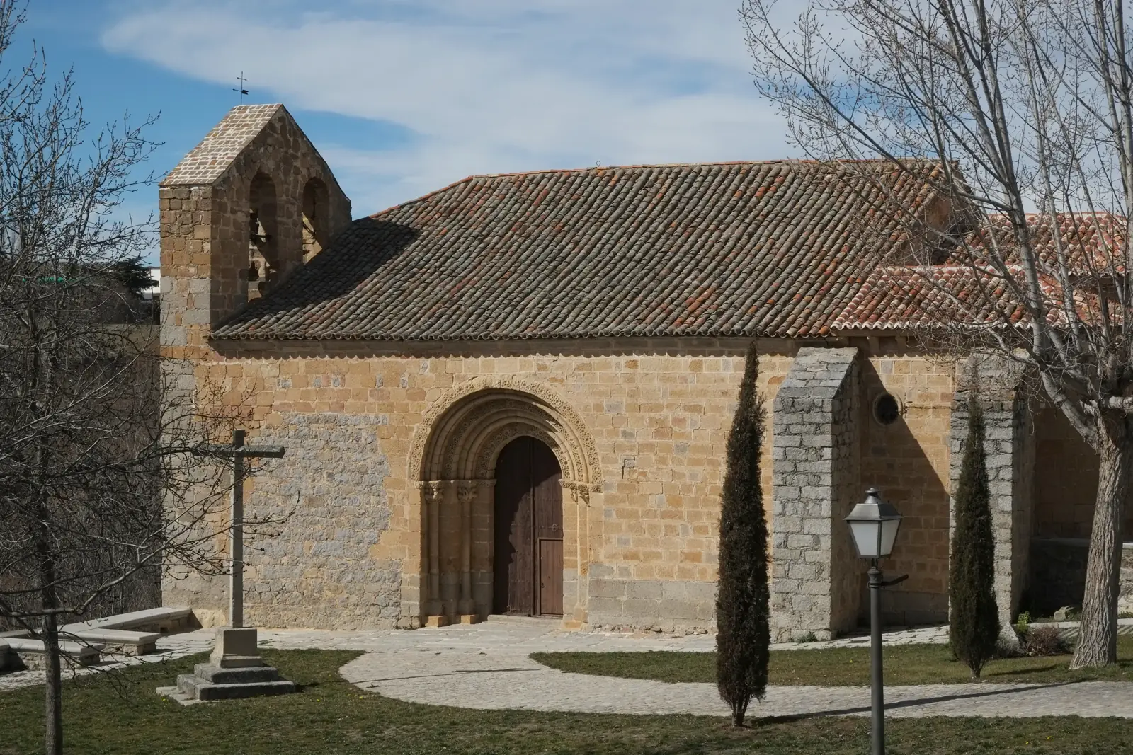 Fachada sur de la ermita de San Segundo de Ávila, con portada románica de arquivoltas, espadaña de dos vanos, cruz de piedra y cipreses en el jardín