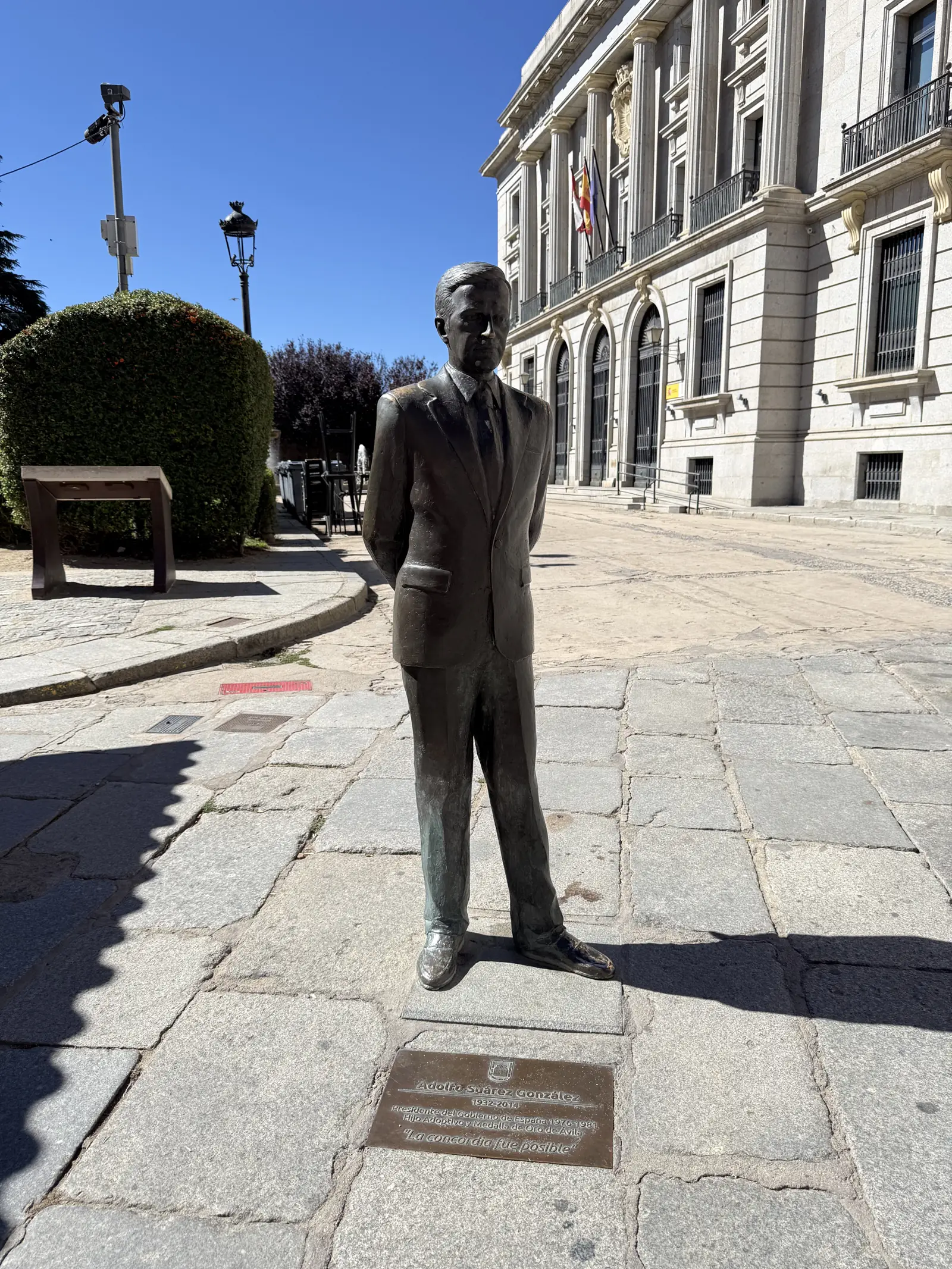 Estatua de bronce de Adolfo Suárez de pie sobre el enlosado ante un edificio neoclásico con banderas en Ávila. Foto propia La Maleta Inquieta, septiembre 2026