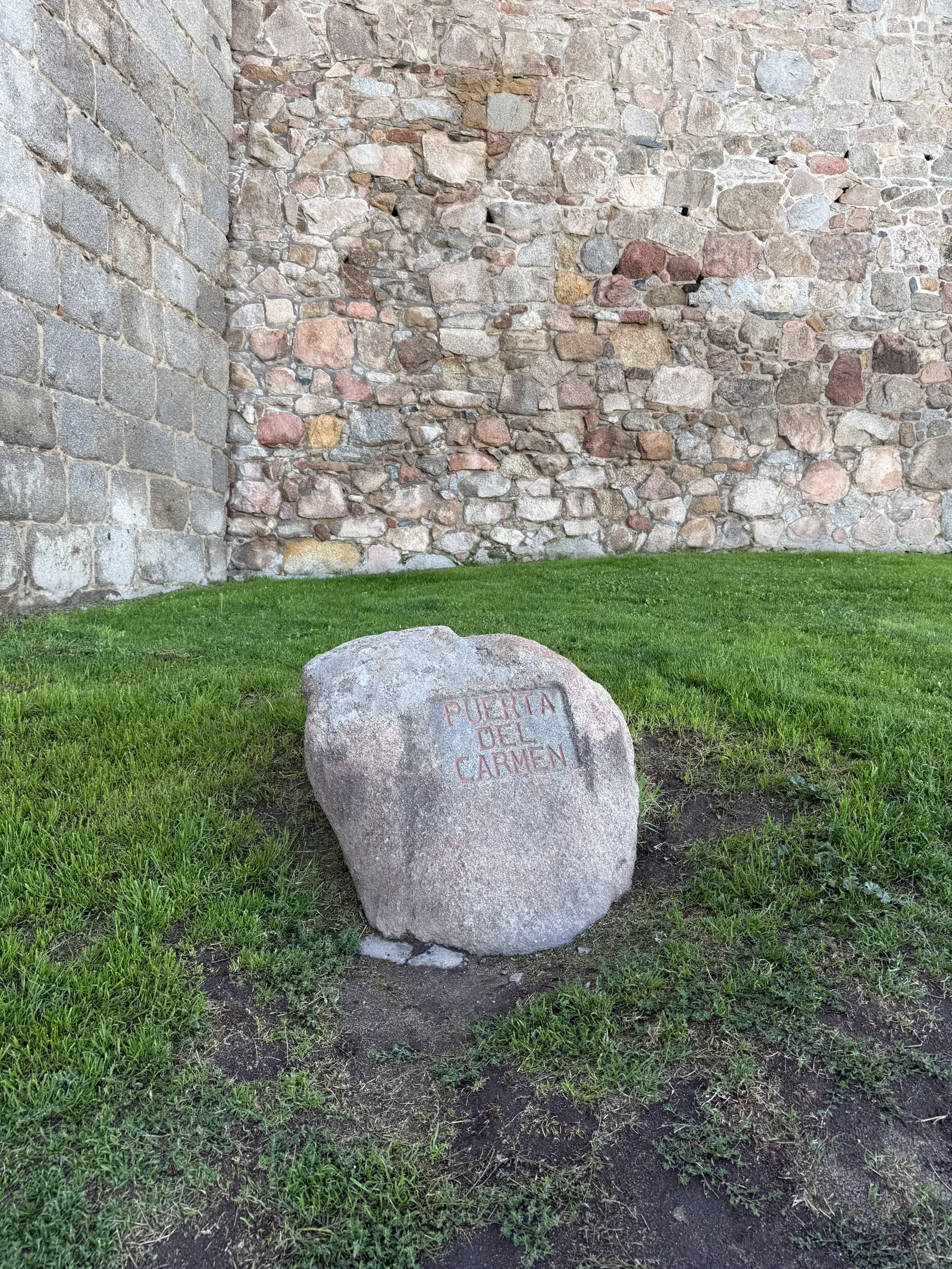 Bolo de granito con Puerta del Carmen grabado en rojo sobre el césped, al pie de la muralla norte de Ávila. Foto propia La Maleta Inquieta, septiembre 2026.