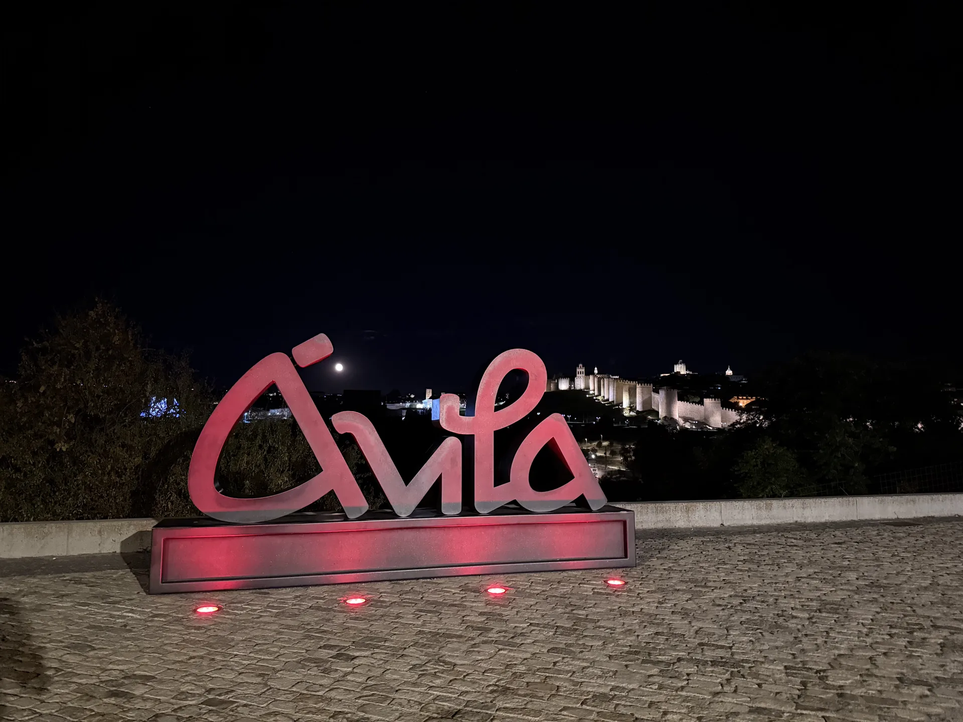 Letras de Ávila iluminadas en rojo de noche en Los Cuatro Postes, con la luna y la muralla iluminada al fondo. Foto propia La Maleta Inquieta, septiembre 2026.