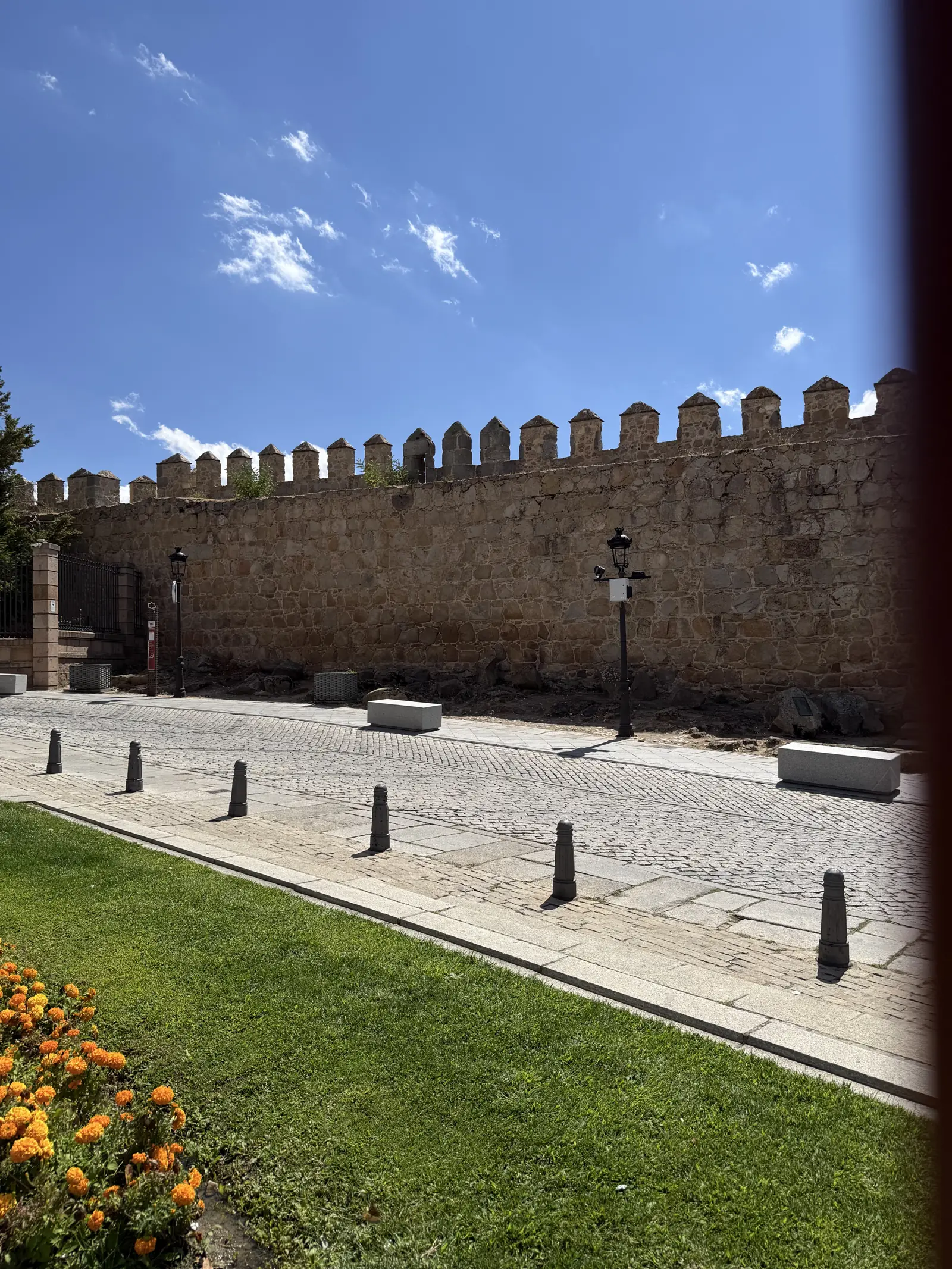 Lienzo recto y almenado de la muralla de Ávila junto a la plaza de La Santa, con bolardos, césped y flores. Foto propia La Maleta Inquieta, septiembre 2026.
