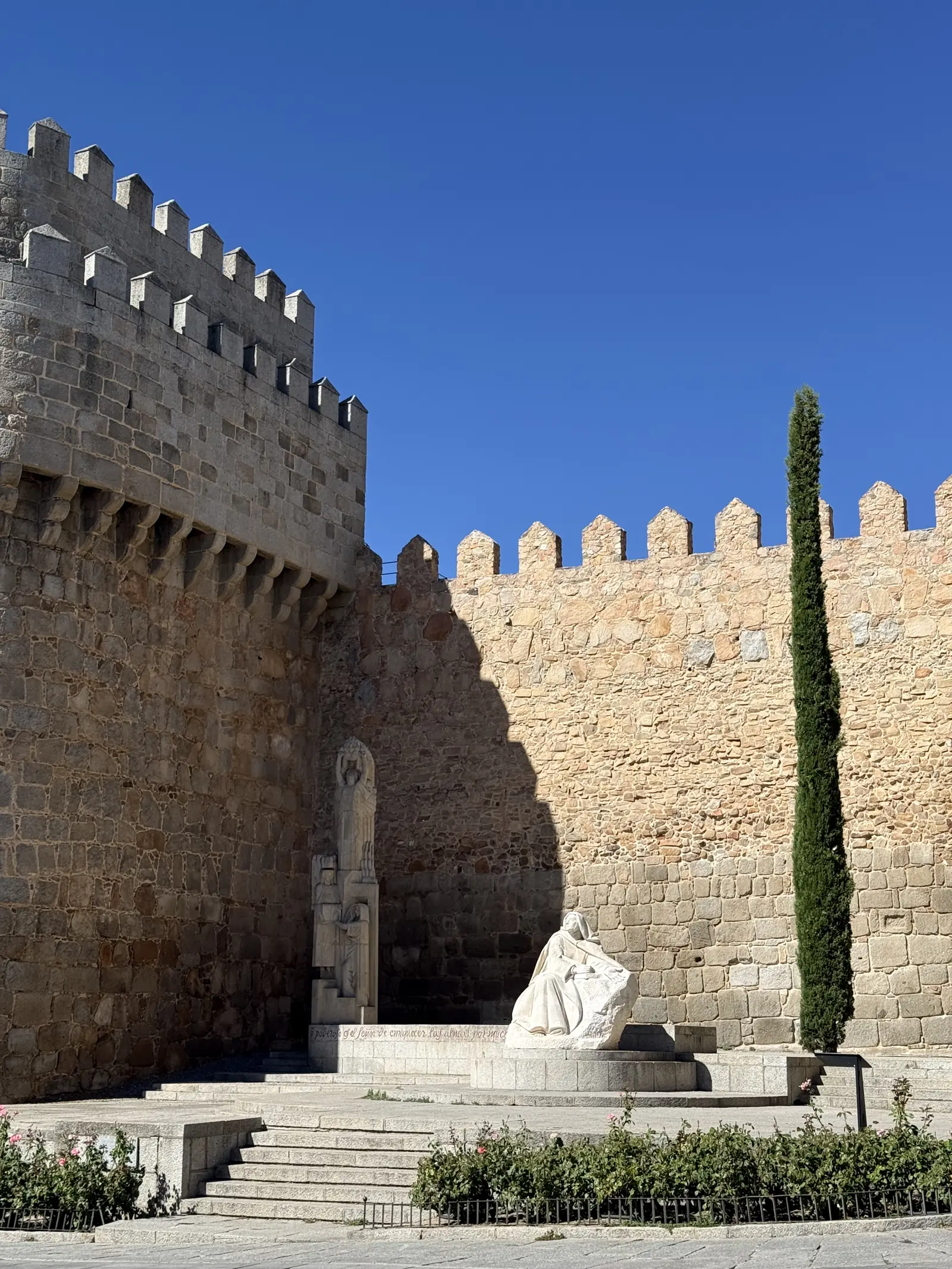Monumento a Santa Teresa sentada ante la muralla en la plaza de Santa Teresa de Ávila, junto a un ciprés. Foto propia La Maleta Inquieta, septiembre 2026