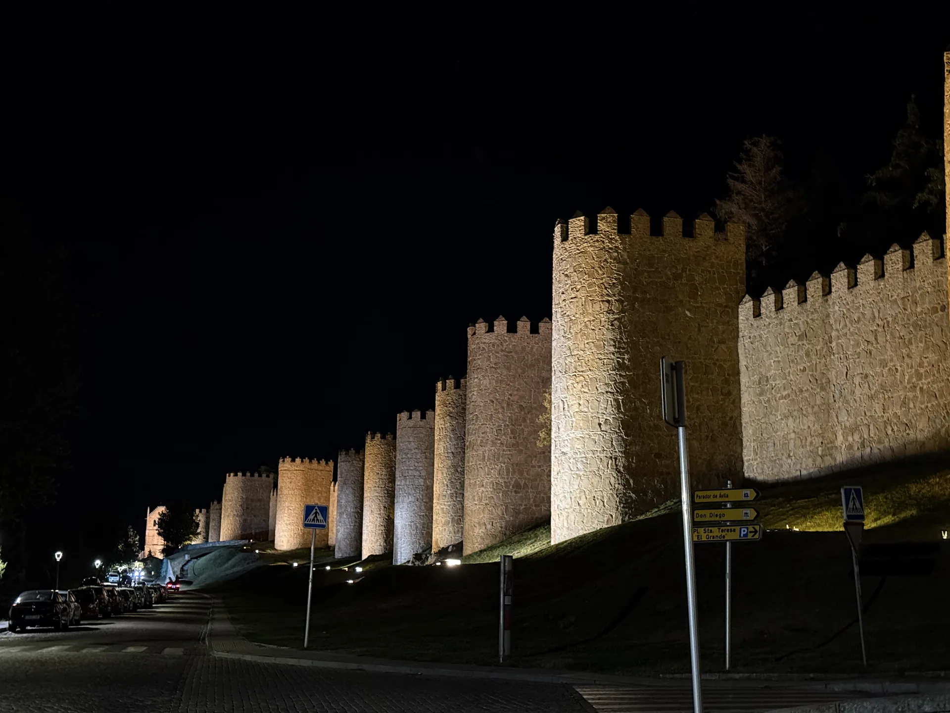 Cubos del lienzo norte de la muralla de Ávila iluminados de noche junto a la calle, con señales hacia el Parador. Foto propia La Maleta Inquieta, septiembre 2026.