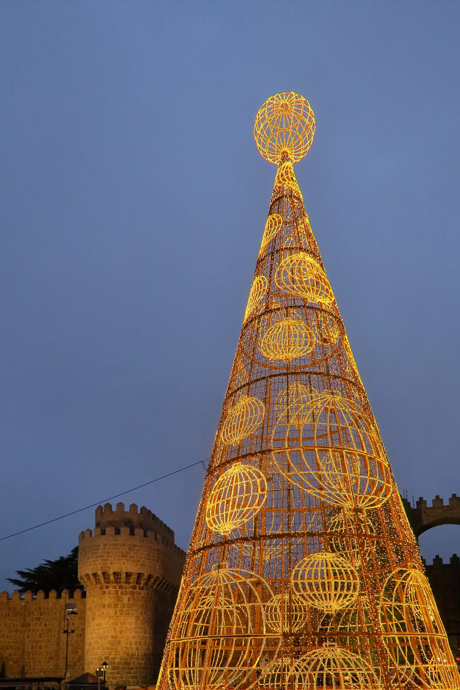Árbol de Navidad de luces doradas en la plaza de Santa Teresa de Ávila, con los cubos de la muralla detrás al anochecer