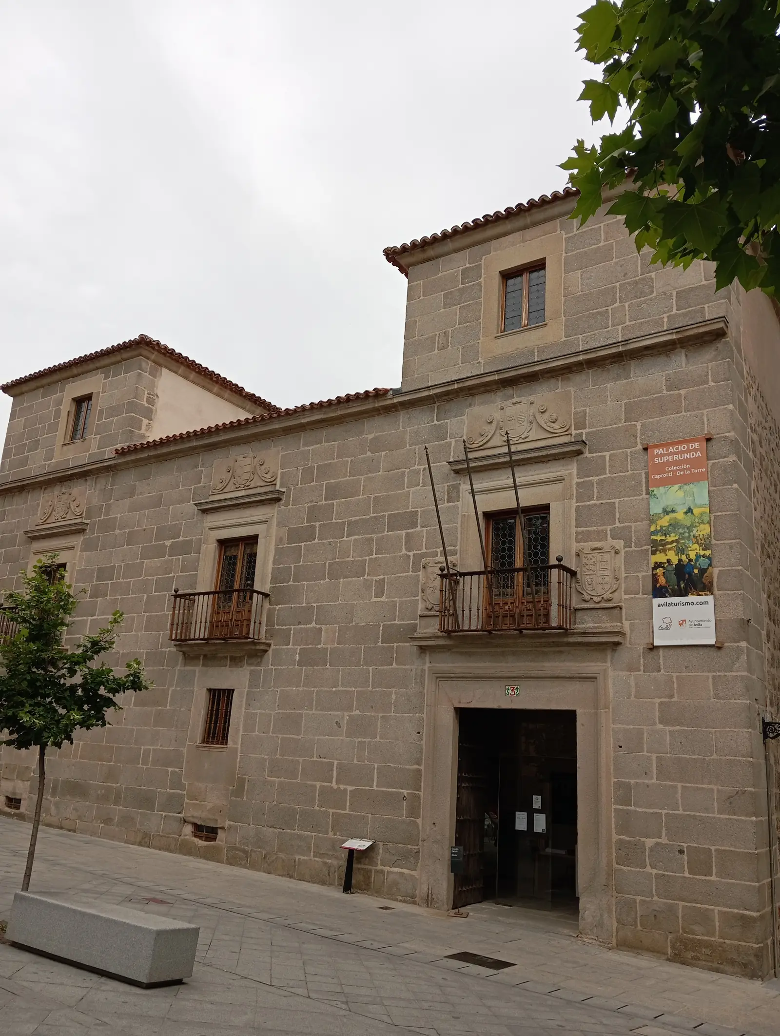 Fachada de granito del palacio de Superunda de Ávila, con escudos, balcones de hierro, dos torres de tejado y el cartel de la colección Caprotti