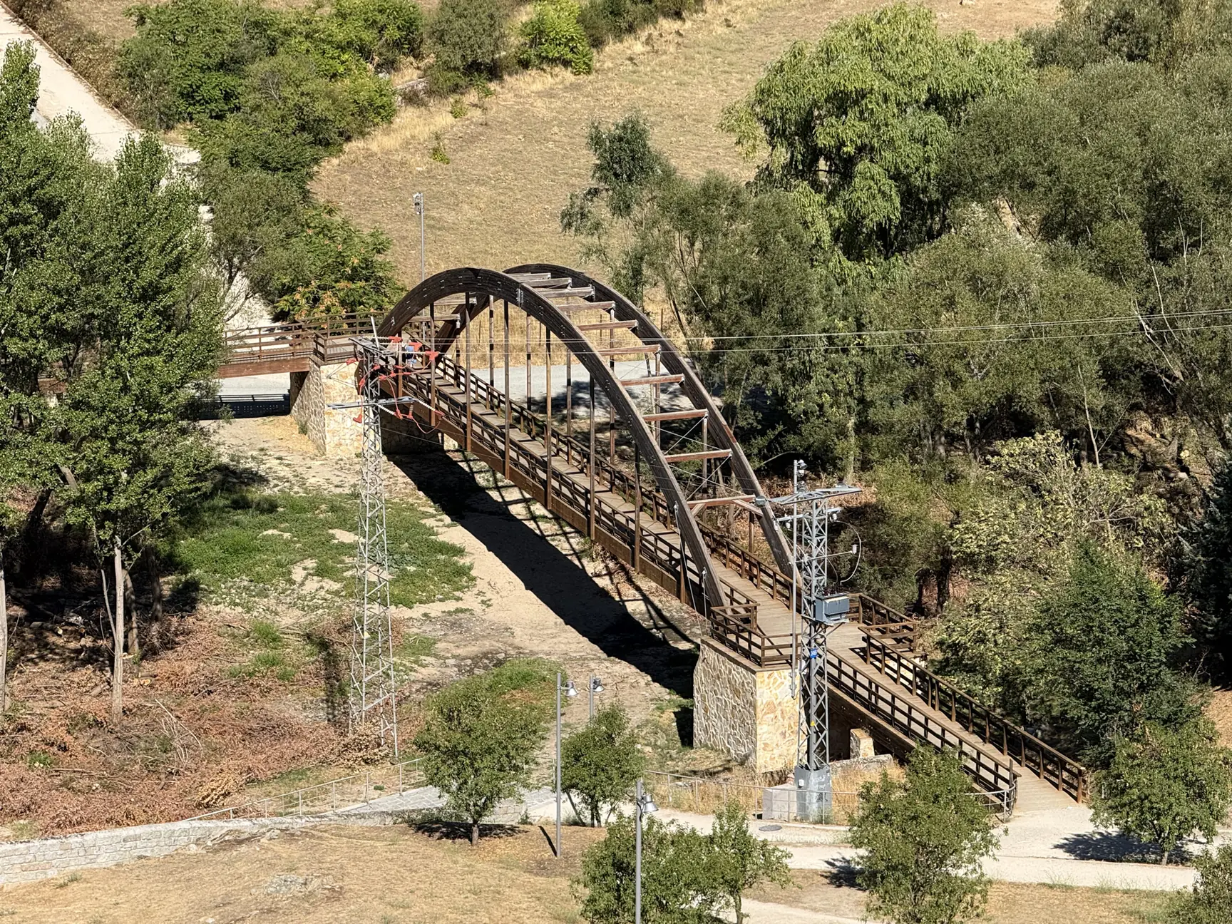 Pasarela peatonal de madera en arco sobre el río Adaja en Ávila, entre encinas y taludes secos. Foto propia La Maleta Inquieta, septiembre 2026.