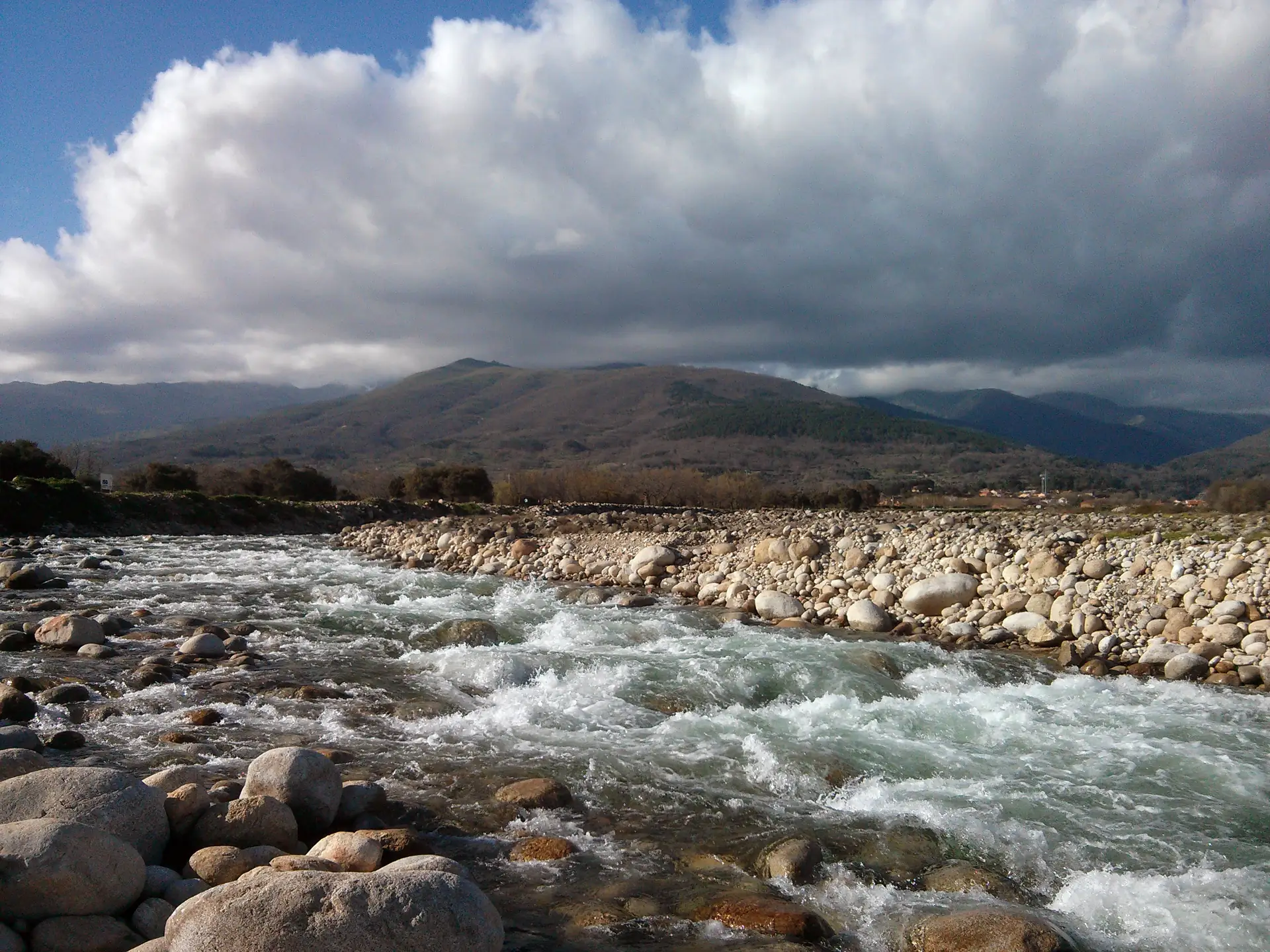 Garganta de Santa María en Candeleda, con el agua corriendo entre cantos rodados y la Sierra de Gredos bajo nubes al fondo