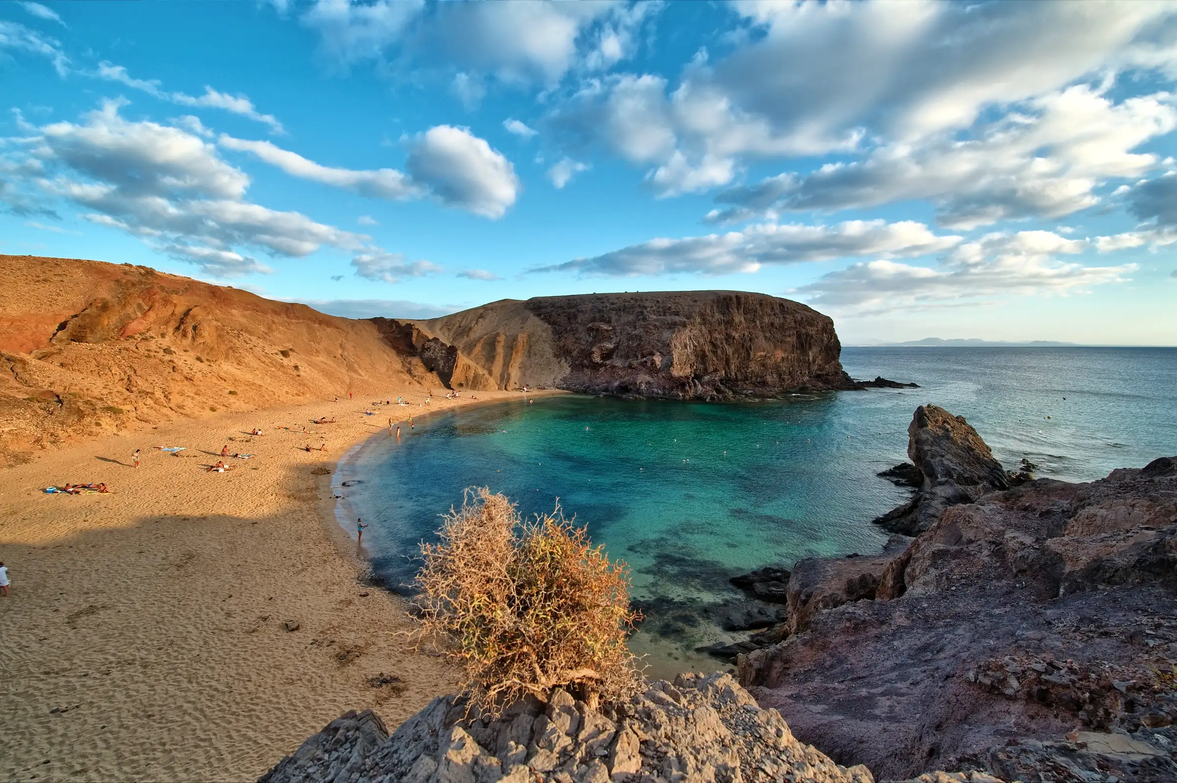 Playa de Papagayo en Lanzarote, con el agua turquesa, gente en la arena y los acantilados rojizos al fondo