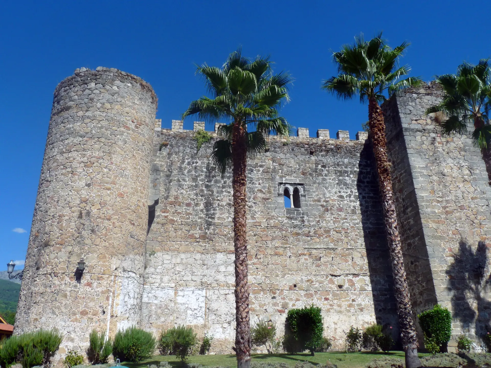 Muralla almenada y torre cilíndrica del castillo de la Triste Condesa en Arenas de San Pedro, con palmeras delante y cielo azul intenso