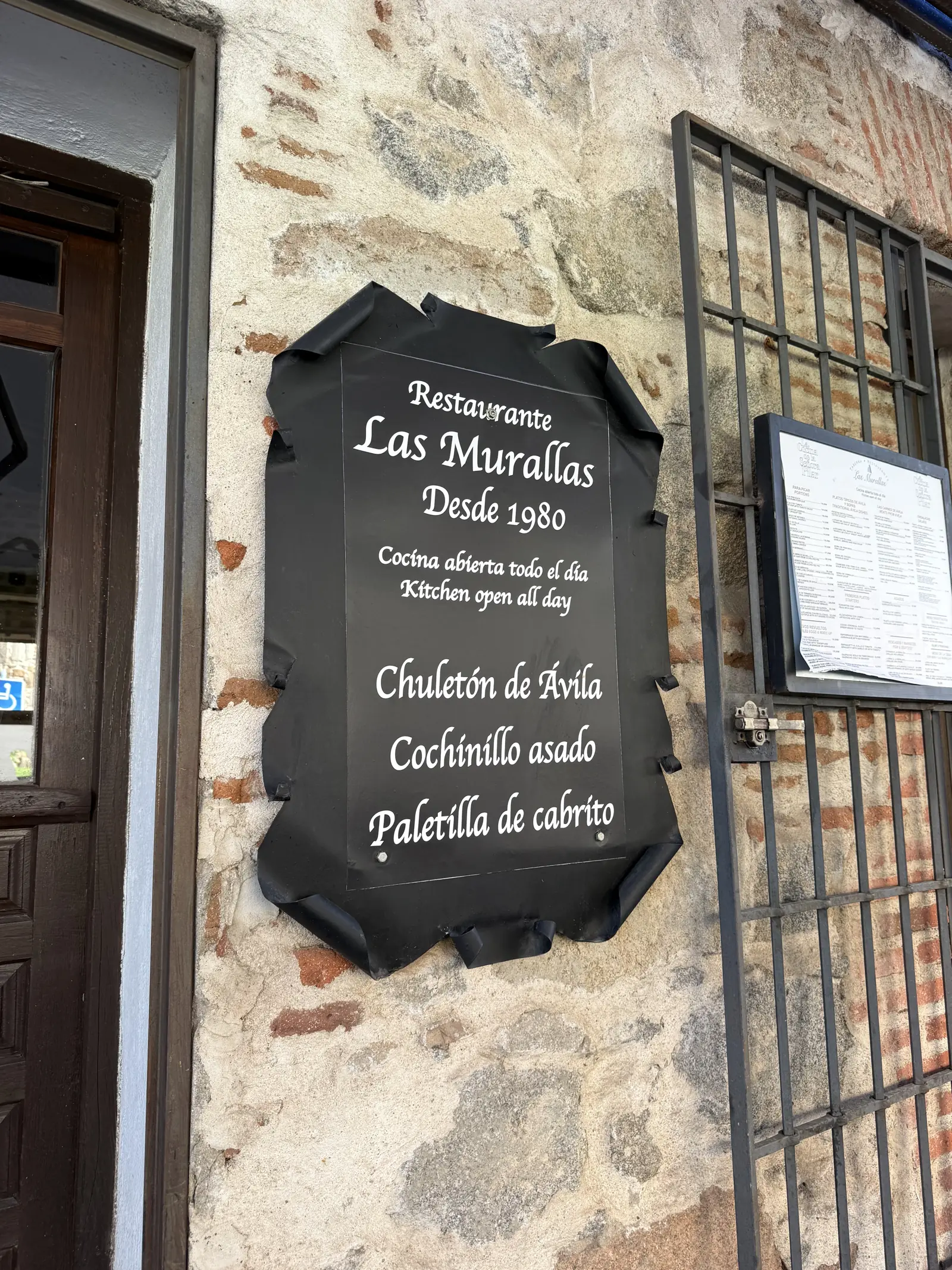 Cartel del restaurante Las Murallas de Ávila con chuletón, cochinillo y cabrito, junto a su carta tras una reja. Foto propia La Maleta Inquieta, septiembre 2026