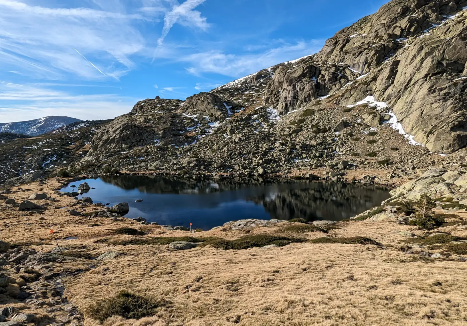Laguna Grande de Peñalara entre prados secos y laderas rocosas con restos de nieve, en la Sierra de Guadarrama