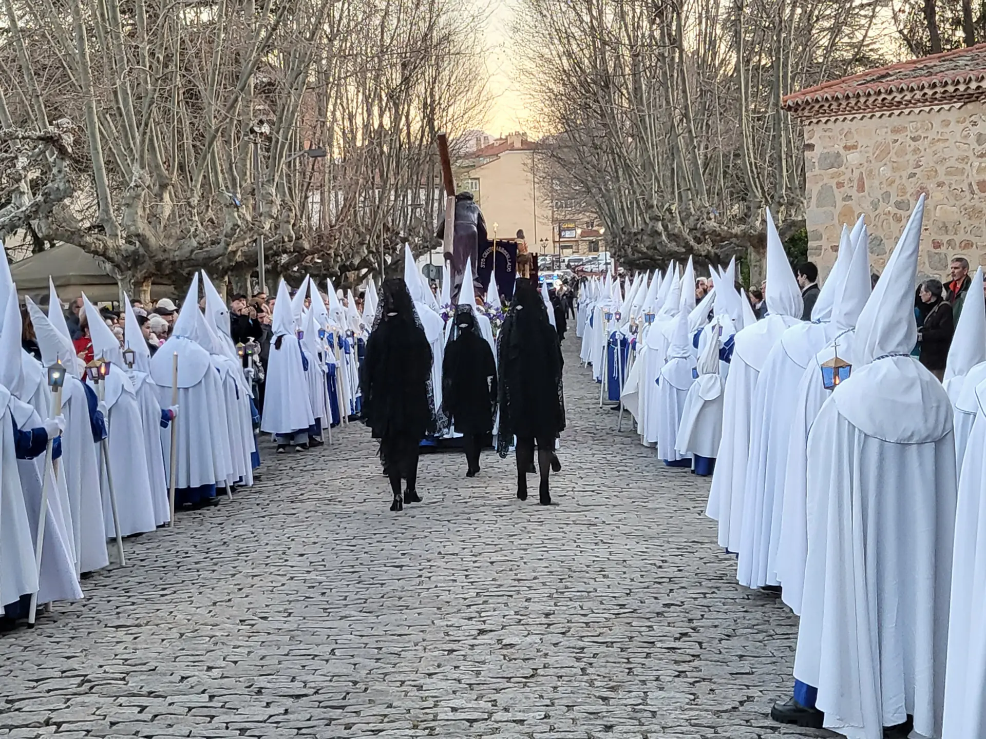 Nazarenos con capirote blanco en dos filas por una calle empedrada junto a la iglesia de San Nicolás en la Semana Santa de Ávila