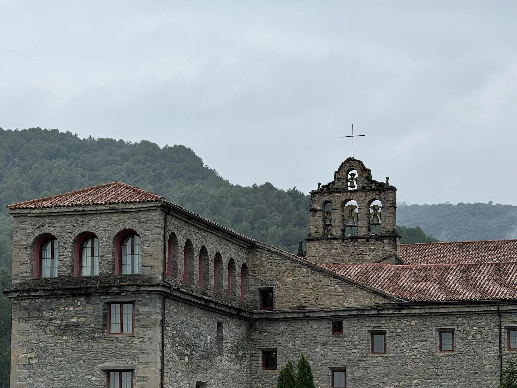 Espadaña de piedra con cuatro campanas y cruz del Monasterio de Boltaña, con su galería de arcos y montes de pinos al fondo. Foto propia La Maleta Inquieta, octubre 2026