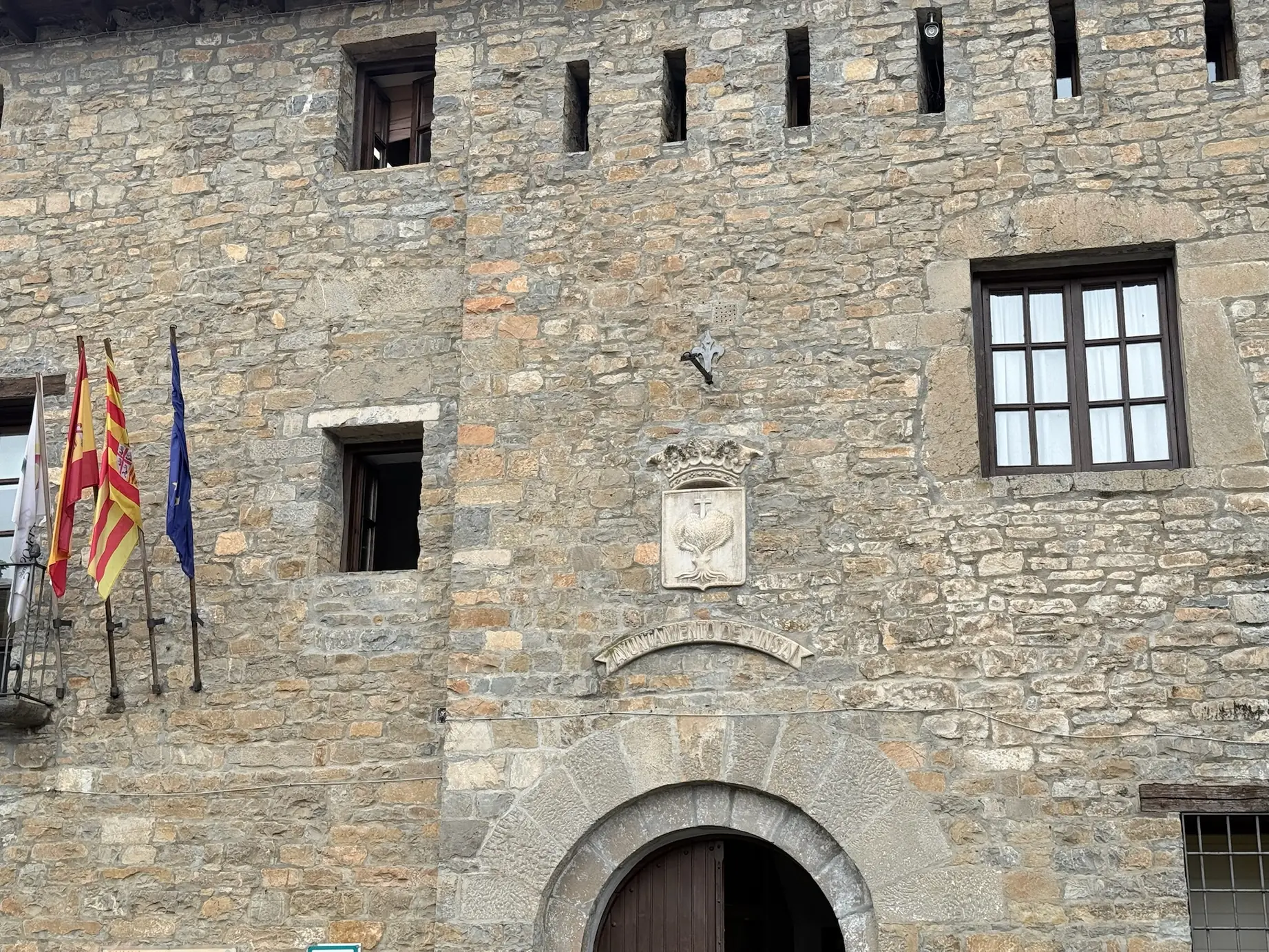 Fachada de piedra del Ayuntamiento de Aínsa con arco de medio punto, escudo de la Cruz de Sobrarbe y banderas de España, Aragón y la UE. Foto propia La Maleta Inquieta, octubre 2026
