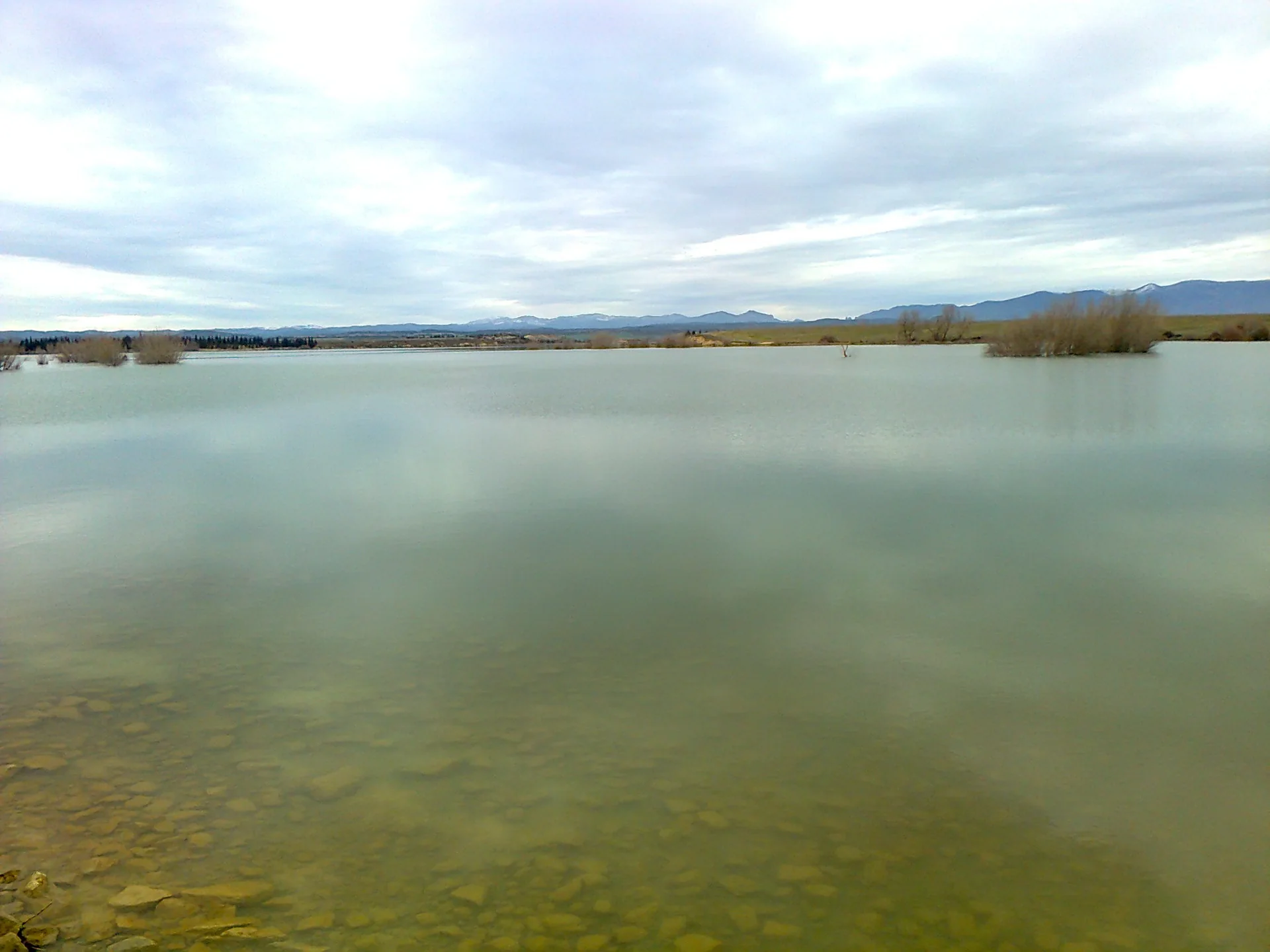 Agua tranquila del embalse de la Sotonera desde su orilla llana, con la sierra prepirenaica en el horizonte