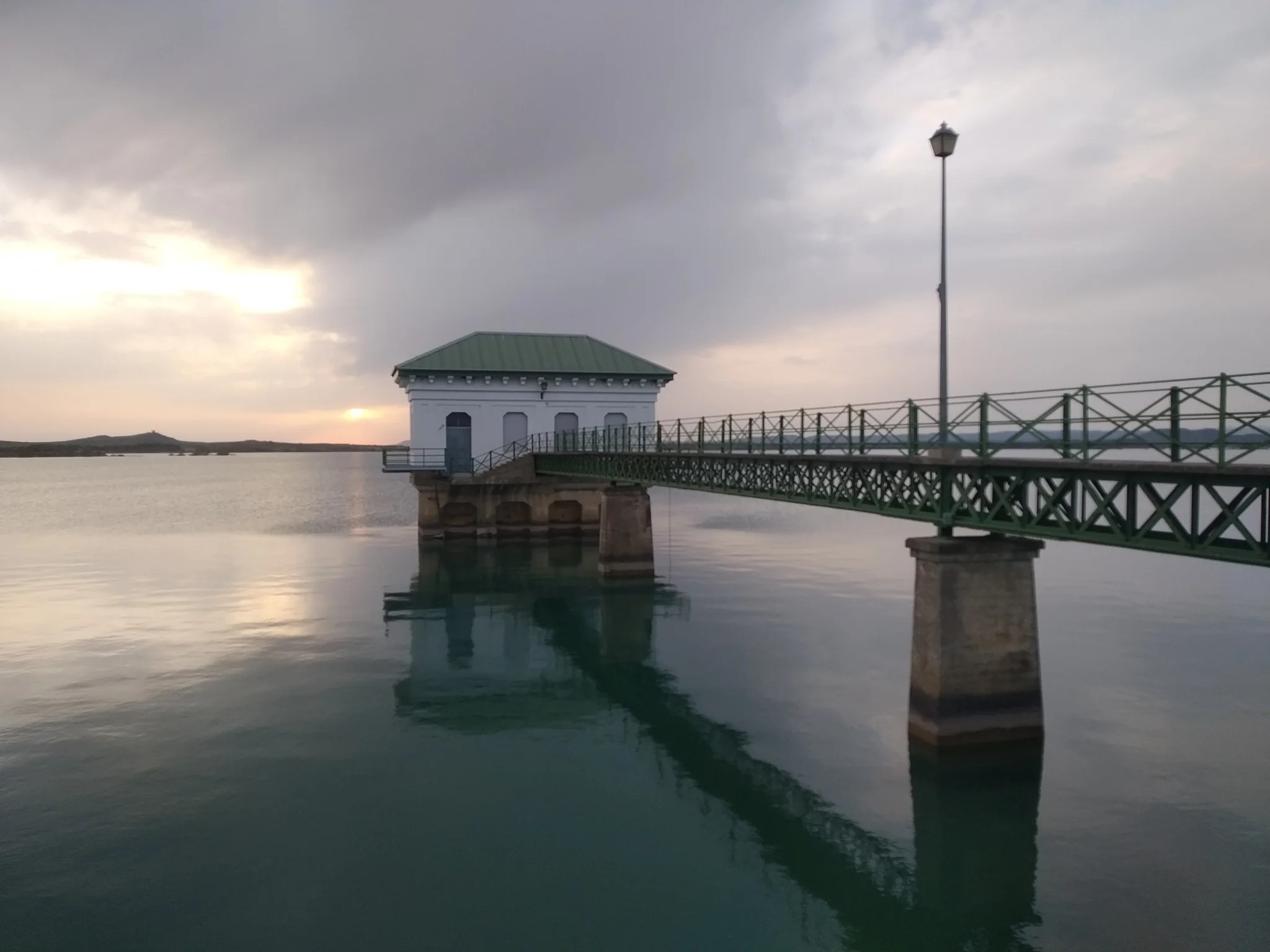 Embalse de La Sotonera con pasarela metálica y caseta sobre el agua al atardecer bajo cielo nublado con reflejos dorados