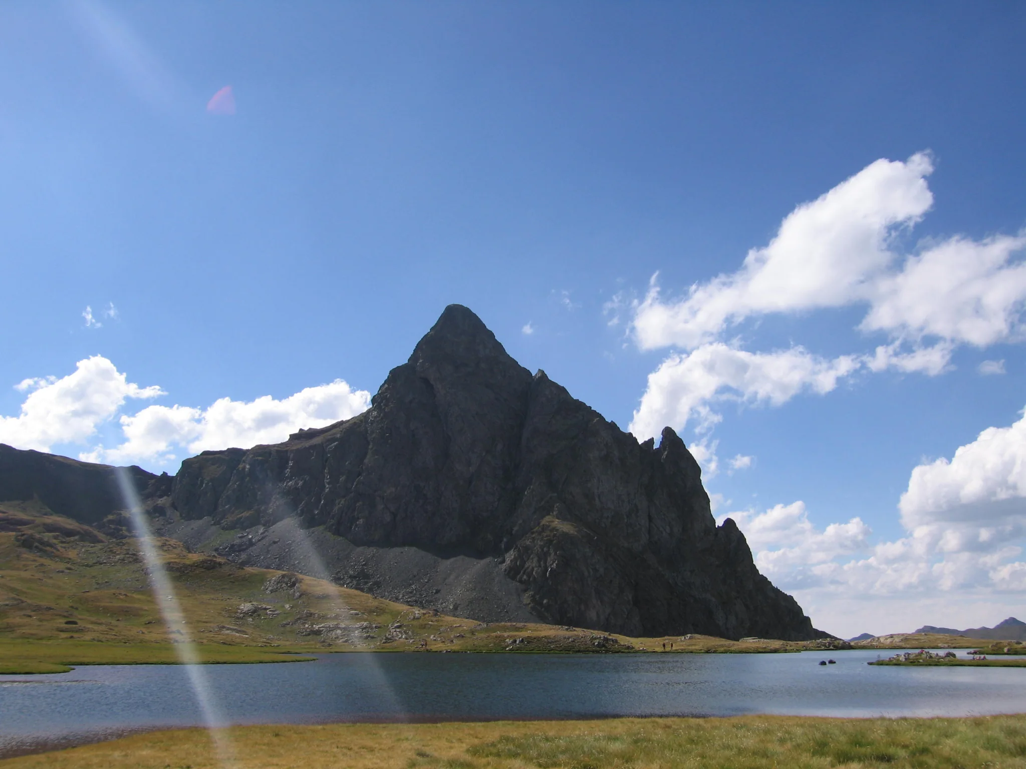 Ibón de Anayet con su característico pico piramidal de roca oscura al fondo y pastos amarillentos en la orilla bajo cielo azul con nubes
