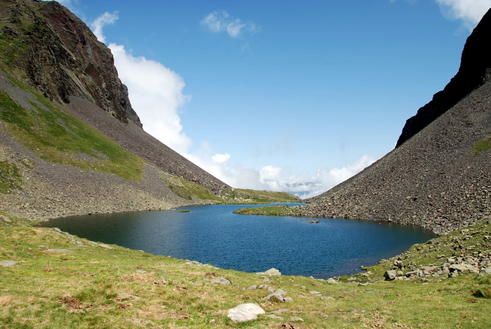 Ibón del Coth del Horo rodeado de laderas pedregosas y prados verdes con cumbres pirenaicas al fondo bajo cielo azul