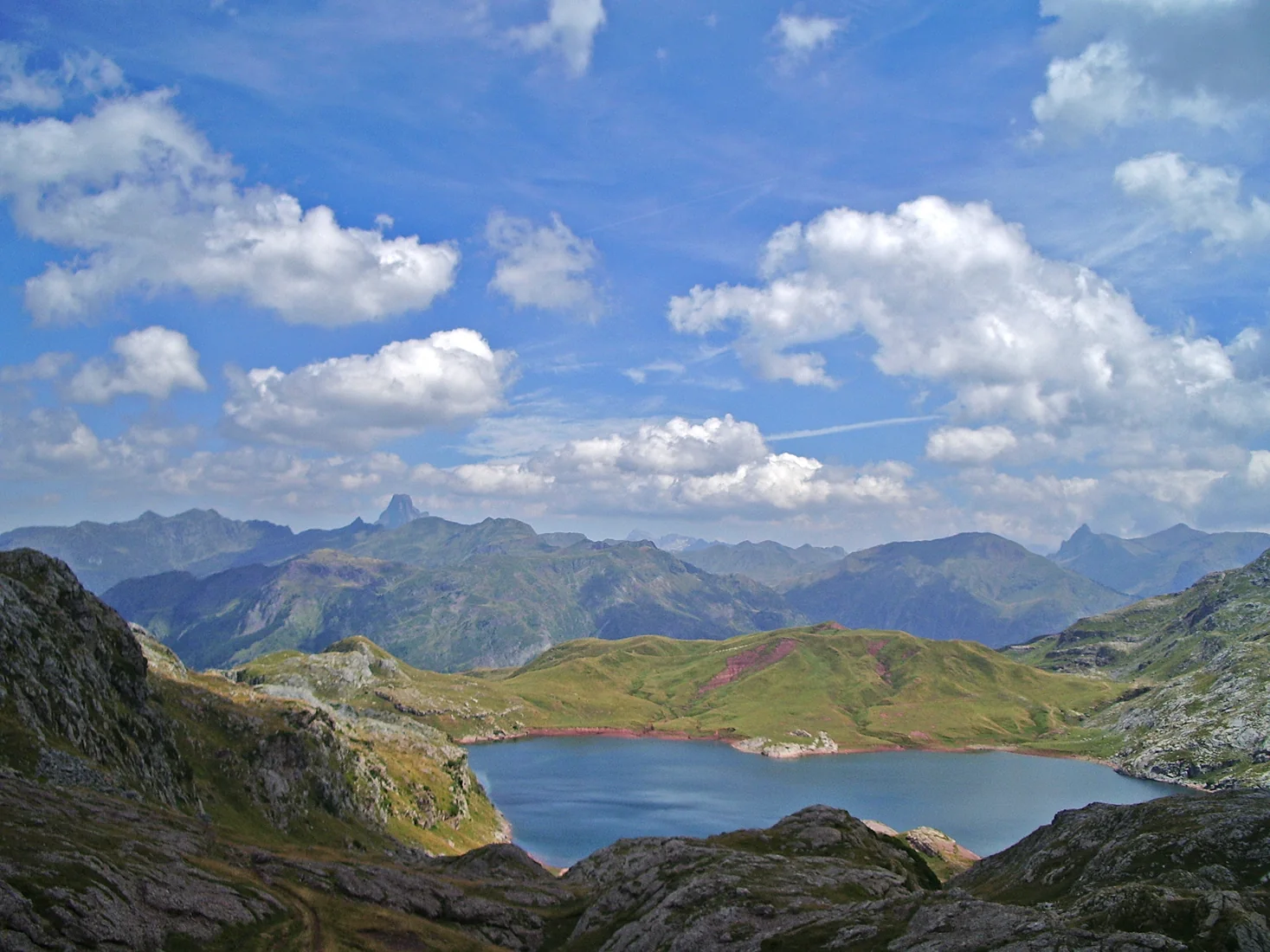 Ibón de Estanés visto desde lo alto, lago azul encajado entre lomas verdes y montañas del Pirineo junto a la frontera