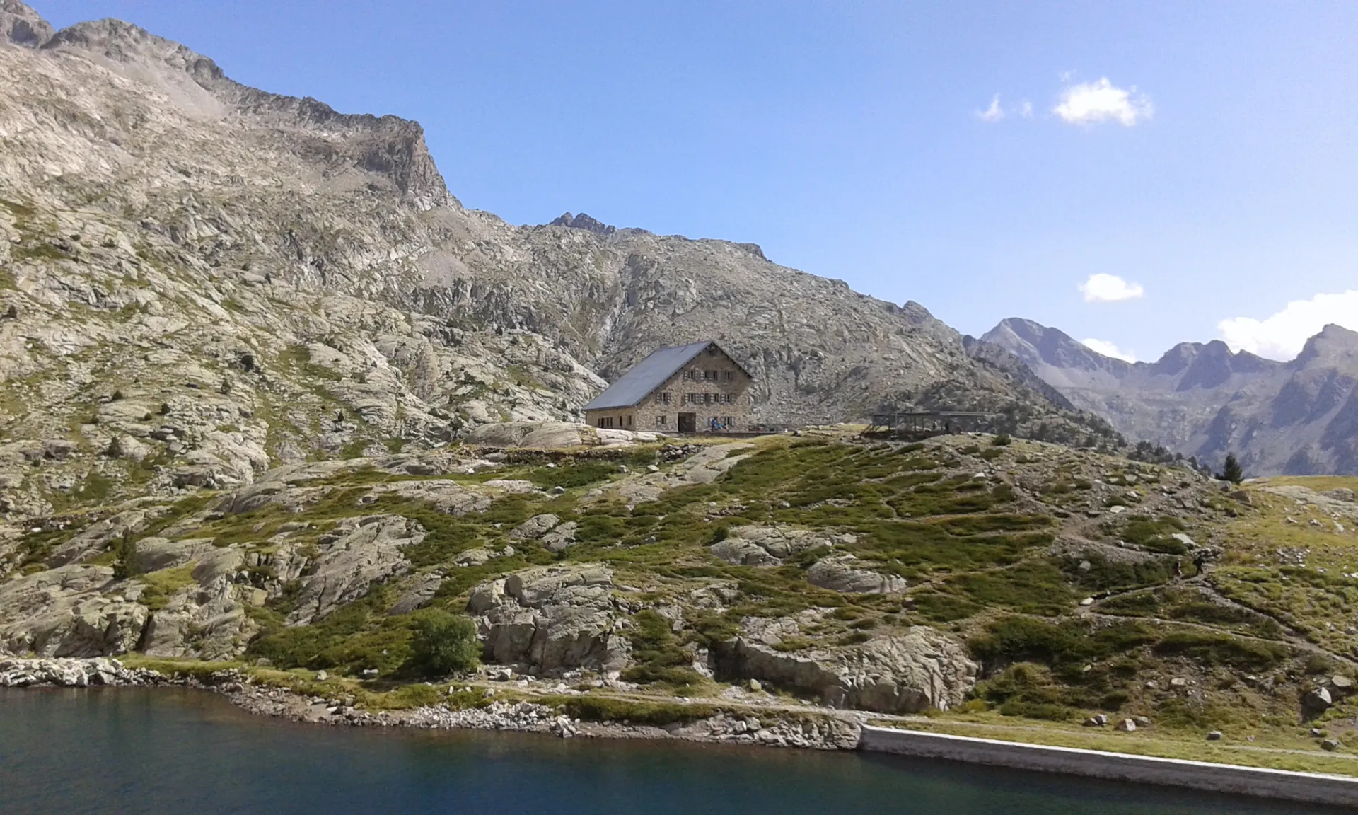 Ibón de Bachimaña con su presa en primer plano y el refugio de piedra sobre la ladera granítica, en la alta montaña