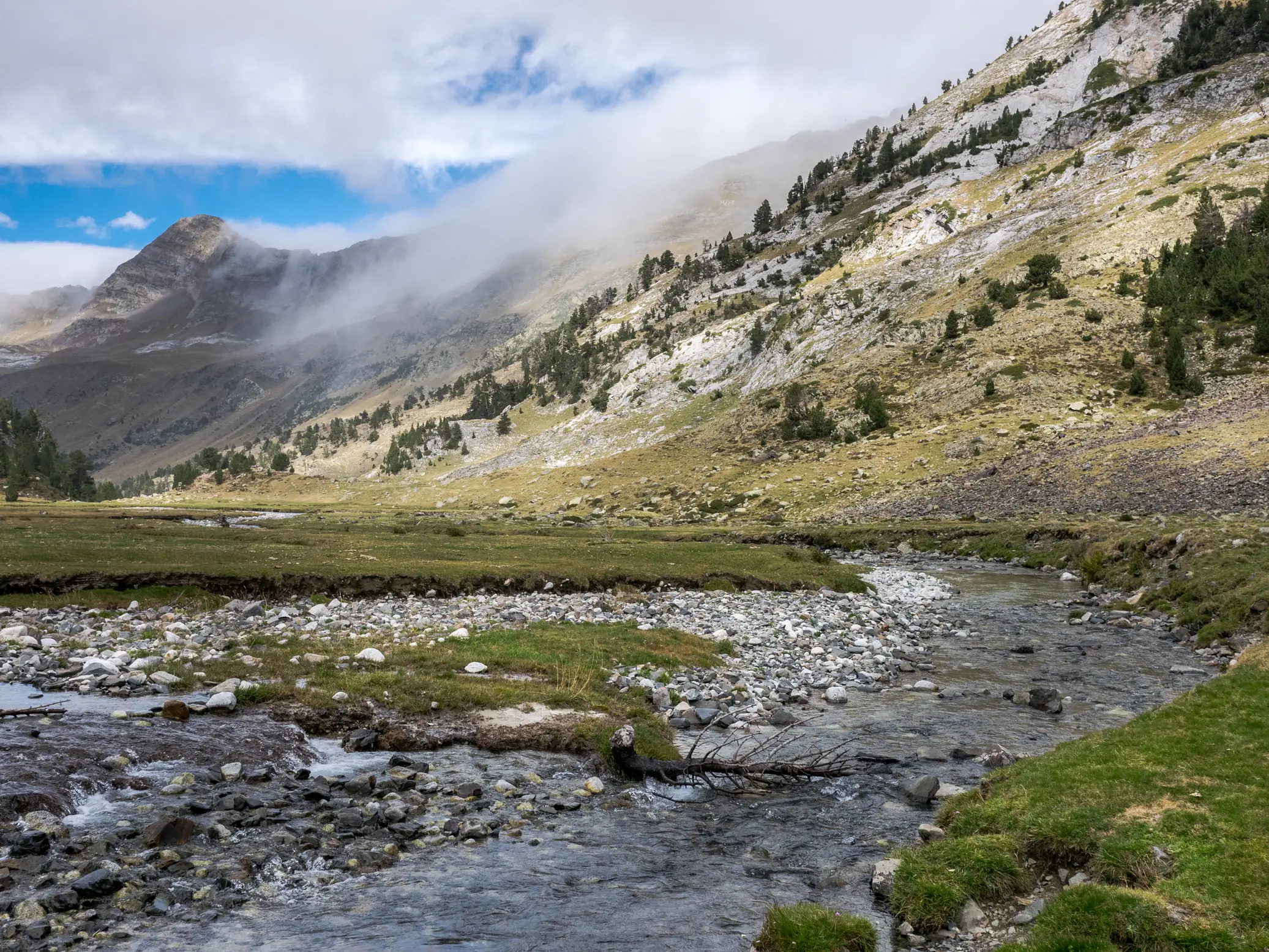 Paisaje del llano de Aigualluts en Benasque con río de montaña entre prados verdes y laderas con niebla dispersa