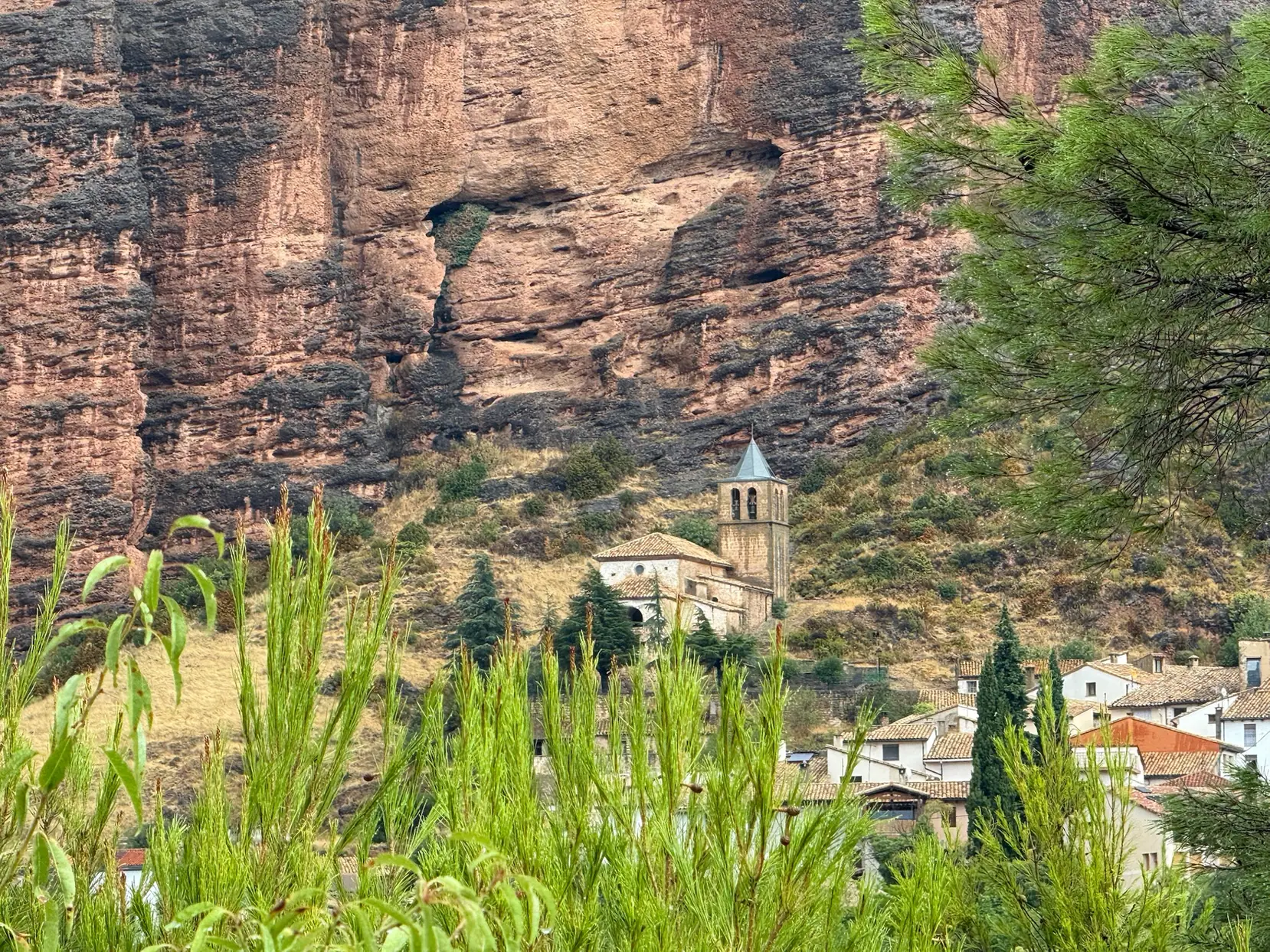 Iglesia de Riglos entre casas y vegetación, bajo la gran pared de conglomerado de los Mallos de Riglos. Foto propia La Maleta Inquieta, octubre 2026